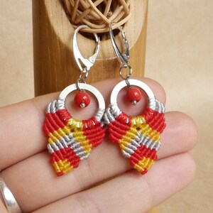 May include: A pair of handmade macrame earrings. The earrings feature a silver hoop with a small red bead and a woven design in red, yellow, and gray. The earrings are displayed against a neutral background.