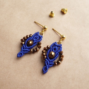 Può includere: Un paio di orecchini in macramè blu con perni e dettagli color oro. Gli orecchini presentano una perla centrale marrone circondata da perline marroni più piccole. Sono visibili anche due chiusure per orecchini dorate.
