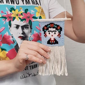 May include: A small, woven wall hanging featuring a pixelated portrait of Frida Kahlo. The artwork is in shades of blue, black, red, and beige, with a fringe at the bottom. The piece is suspended from a wooden dowel with a white cord.