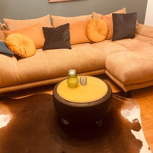 Könnte beinhalten: Ein brauner Kuhfellteppich mit einem Reifen-Couchtisch mit einer Holzplatte. Der Tisch hat eine grüne Glasvase und einen Kerzenhalter aus Metall darauf. Der Tisch steht vor einem braunen Ecksofa mit mehreren Kissen.