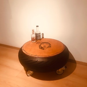 Könnte beinhalten: Ein umgebauter Reifen als Beistelltisch mit einer Holzplatte. Die Platte hat eine gealterte Holzoptik und eine schwarz-weiße Grafik eines Frauenkopfes. Zwei Flaschen Alkohol stehen auf dem Tisch.