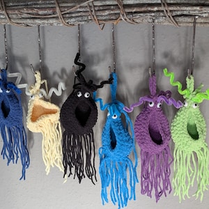 Könnte beinhalten: Eine Reihe handgefertigter, skurriler Häkeltiere in verschiedenen Farben, darunter Blau, Gelb, Schwarz und Lila, jedes mit Wackelaugen und hängenden Fransen. Sie hängen an Metallhaken, die an einem rustikalen Ast befestigt sind.
