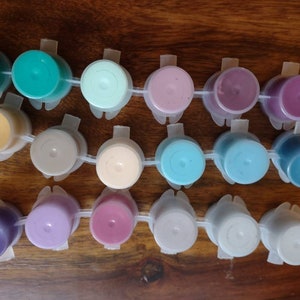 Op de afbeelding: Een set van 16 kleine verfpotjes met deksels, georganiseerd in een plastic bakje. De verfkleuren omvatten teal, lichtgroen, roze, paars, geel, beige, lichtbruin, lichtblauw, grijs en wit.