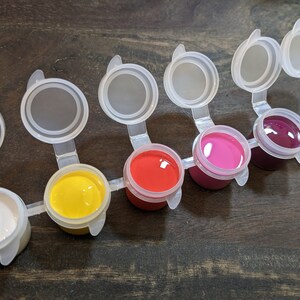 Könnte beinhalten: Ein Set aus sechs kleinen Farbtöpfen mit verschiedenen Farben, darunter Weiß, Gelb, Rot, Rosa, Burgunder und Lila. Jeder Topf hat einen transparenten Kunststoffdeckel. Die Farbtöpfe sind auf einer dunklen Holzoberfläche angeordnet.