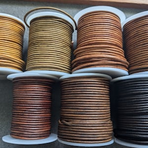 Könnte beinhalten: Spulen mit Lederkordel in verschiedenen Farben, darunter Braun, Beige und Schwarz. Die Kordeln sind um weiße Spulen gewickelt.
