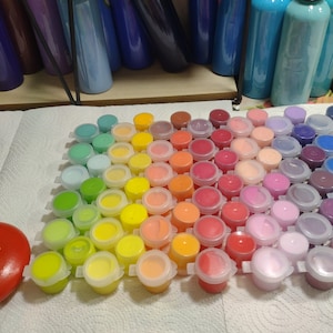 Op de afbeelding: Een set van 50 kleine verfpotjes, gerangschikt in een regenboogpatroon. De potjes zijn van helder plastic met witte deksels en bevatten verschillende kleuren acrylverf.