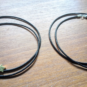 Könnte beinhalten: Zwei Halsketten mit schwarzen Kordeln und Sternanhängern. Eine Halskette hat einen goldfarbenen Stern und Verschluss, die andere einen silberfarbenen Stern und Verschluss. Die Halsketten liegen auf einer Holzoberfläche.