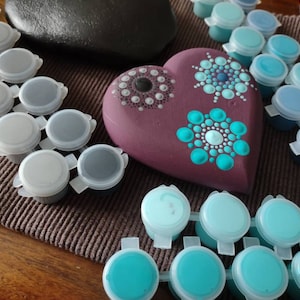 May include: A heart-shaped purple stone with three turquoise mandala designs painted on it.  Surrounding the stone are multiple small containers of turquoise paint.