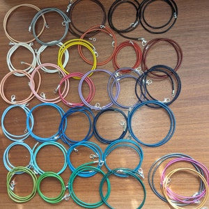 Op de afbeelding: Een verzameling van 20 lederen koordsnoeren in verschillende kleuren, waaronder zwart, bruin, blauw, groen en rood. De snoeren zijn allemaal ongeveer even lang en breed, en ze zijn in een cirkelvormige opstelling op een houten oppervlak gerangschikt.