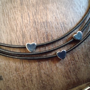 Peut inclure: Gros plan sur un collier avec trois brins de cordon noir. Chaque brin comporte un petit charme en forme de cœur argenté. Le collier est présenté sur une surface en bois.