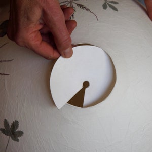 May include: A white paper lampshade with a round hole cut out. A white paper circle with a small hole in the center is being held over the hole in the lampshade.