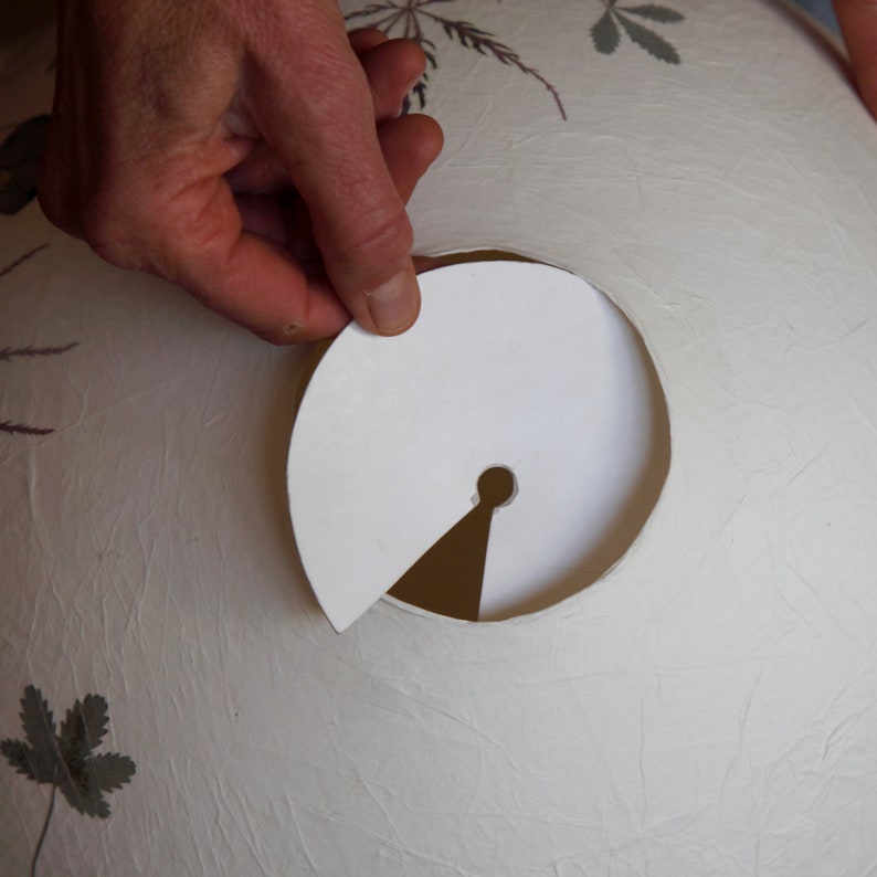 May include: A white paper lampshade with a round hole cut out. A white paper circle with a small hole in the center is being held over the hole in the lampshade.