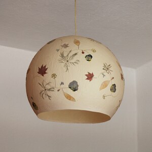 May include: A round, cream-colored lampshade with a floral design made from pressed flowers. The lampshade is hanging from a white ceiling.