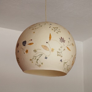 May include: A white round lampshade with dried flowers and leaves pressed into the surface. The lampshade is hanging from a white ceiling.