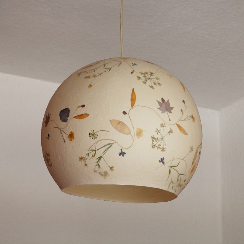 May include: A white round lampshade with dried flowers and leaves pressed into the surface. The lampshade is hanging from a white ceiling.