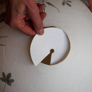 May include: A white paper lampshade with a circular opening. A hand is holding a white paper template with a small hole in the centre, which is being used to cut out the opening in the lampshade.