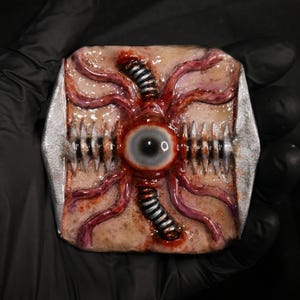 May include: A grotesque, rectangular prop with a central eye, surrounded by red veins and metallic accents. The design includes rows of sharp teeth and a stitched, mechanical element, creating a horror-themed aesthetic.