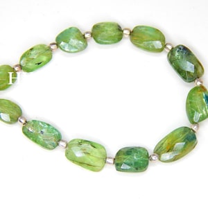 May include: A bracelet featuring faceted, oval-shaped green gemstones. The stones are connected by small, silver-colored beads. The bracelet is displayed on a white surface, showcasing the vibrant green color of the stones.