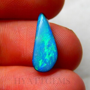 May include: A teardrop-shaped opal gemstone, displaying a vibrant play of color. The stone features a base of deep blue with flashes of bright green and turquoise. The text "HYATT GEMS" is visible at the bottom.