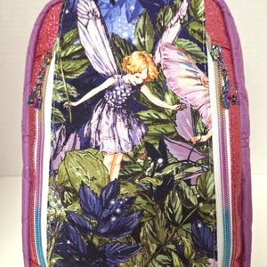 May include: A small, purple backpack with a fairy and butterfly design. The front panel features a colourful illustration of a fairy in a forest setting. The backpack has a glittery red trim and two zippered compartments with decorative pulls.