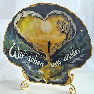 Könnte beinhalten: Handbemalte Muschelkunst mit einer herzförmigen Landschaft, in der eine Figur auf eine helle Sonne zugeht. Das Kunstwerk zeigt einen Baum, Berge und den Text "Wir sehen uns wieder..." in Weiß. Die Muschel ist auf einem goldfarbenen Metallständer montiert.