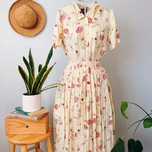 May include: A vintage-style cream-colored dress with a floral pattern of pink roses. The dress features short sleeves, a collared neckline, and a pleated skirt. A straw hat hangs on the wall, and a wooden stool with a plant and books is in the foreground.