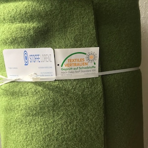 May include: A roll of green fabric, tightly bound with a white string. The fabric has a textured appearance. Attached tags display the text "STOFFE LORENZ" and "TEXTILES VERTRAUEN".