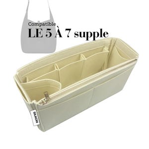 May include: Cream-colored bag organizer with multiple compartments and a zippered closure. The organizer is designed to fit inside a handbag and features several pockets for organization. The text "Compatible LE 5 À 7 supple" is visible.