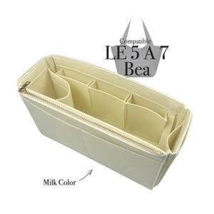 Puede incluir: Un organizador de bolso de fieltro beige con múltiples compartimentos y cierre de cremallera. El organizador es compatible con el bolso Le 5 A 7 Bea. El texto "Milk Color" está escrito debajo del organizador.