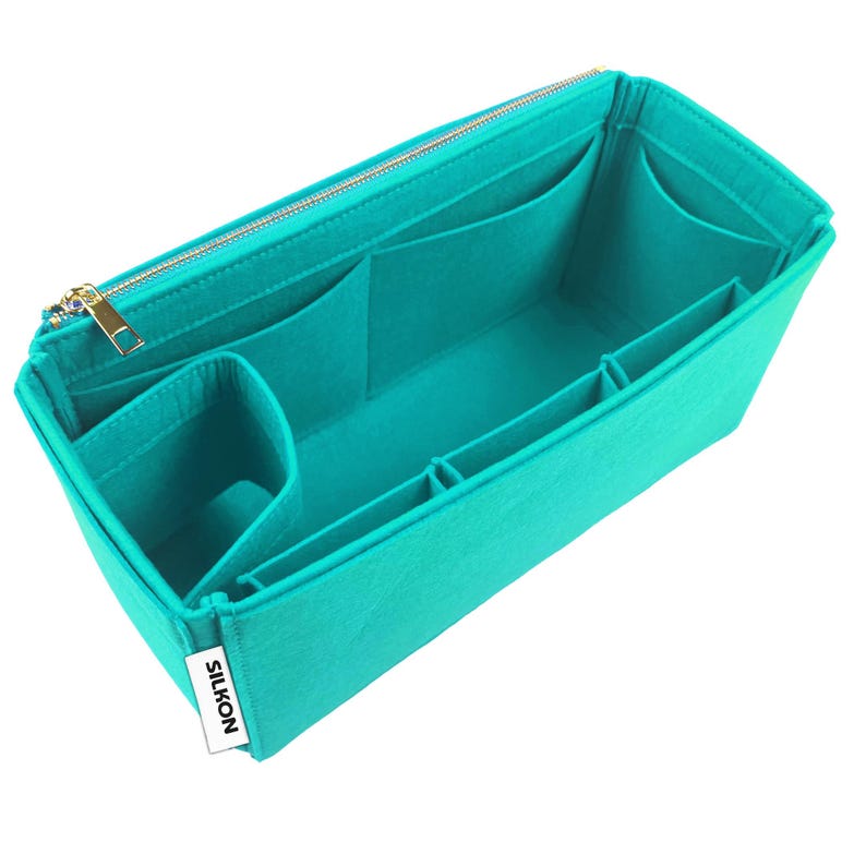 May include: Teal felt bag organizer with multiple compartments and a gold zipper. The organizer has several pockets of varying sizes and a label that reads "SILKON". This insert is designed to help organize the contents of a handbag.