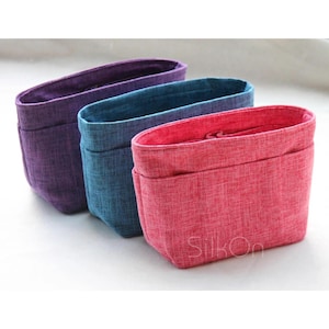 May include: Three fabric organizers in purple, teal, and pink. Each organizer has a rectangular shape with an open top and a front pocket. The organizers are made of a textured material and are designed to help organise items.