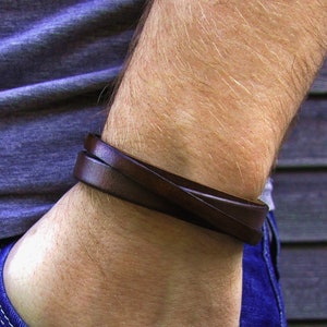 Könnte beinhalten: Ein dunkelbraunes Lederarmband mit einem einfachen Wickeldesign. Das Band besteht aus einem glatten, flachen Lederstreifen, der sich zweimal um das Handgelenk legt und so ein geschichtetes Erscheinungsbild mit sauberen Kanten und einem einheitlichen Farbton erzeugt.