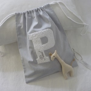 Op de afbeelding: Een witte trekkoordtas met blauwe strepen en een witte viltletter P aan de voorkant. De tas ligt op een wit oppervlak met een klein knuffelgiraffen speelgoed.