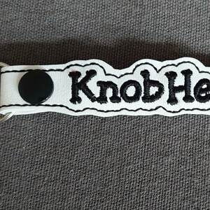 May include: White keychain with the word "KnobHead" embroidered in black. The keychain has a silver clasp and a black button. The design is on a white background with a black border.