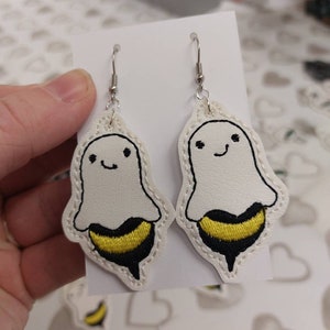 May include: A pair of white ghost earrings with a black and yellow bee design. The earrings are made of felt and have silver hooks.