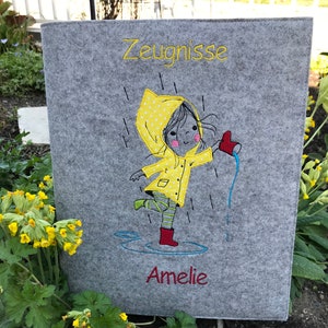 Könnte beinhalten: Grauer Filzordner mit einer Illustration eines Mädchens in einem gelben Regenmantel und dem Text "Zeugnisse" und "Amelie".