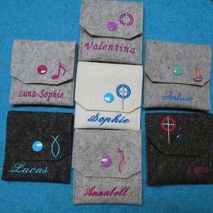 May include: A collection of personalized felt pouches in various colors, each with a button closure and embroidered details. Names like "Valentina" and "Sophie" are embroidered on the pouches, along with decorative symbols. The pouches are arranged on a blue surface.