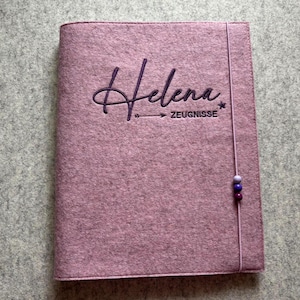Könnte beinhalten: Ein staubrosa Filzcover mit dem Namen "Helena" in dunklerem lila Schriftzug gestickt. Das Wort "ZEUGNISSE" steht unter dem Namen. Ein Band mit drei Perlen sichert das Cover.