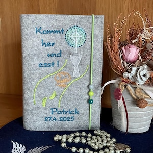 May include: Gray felt book cover with embroidered text "Kommt her und esst!" and a chalice with a cross. The cover also includes the name "Patrick" and the date "27.4.2025".