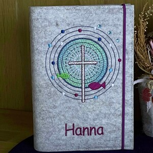 May include: A gray felt book cover with a purple ribbon closure. The cover features an embroidered design of a cross inside a circle with a green and blue background. The name "Hanna" is embroidered in purple at the bottom of the cover.