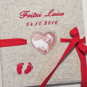 May include: A gray felt photo album with a red ribbon and bow. The album has a heart-shaped window with a photo of a baby inside. The album is embroidered with the name "Fritzi Luise" and the date "04.10.2016".