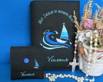 May include: A black felt book cover with blue embroidery of a boat on a wave, the text "Mit Jesus in einem Boot" and the name "Vincent". A smaller black felt pouch with a blue button and the name "Vincent" is also shown. A white rosary with a silver cross is in the foreground.