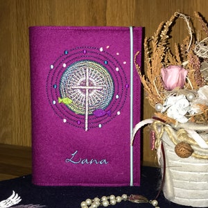 May include: A purple felt book cover with a cross and fish design embroidered in colorful thread. The name "Lana" is embroidered in white thread below the design.