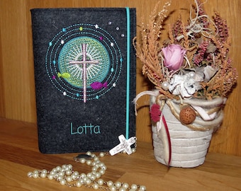 May include: A gray felt book cover with a teal blue trim and an embroidered cross design. The cover has the name "Lotta" embroidered on it. A white pearl rosary with a silver cross pendant is lying on a wooden surface.
