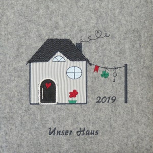Könnte beinhalten: Ein graues Filz-Hauswarming-Schild mit einer gestickten Abbildung eines Hauses, einem roten Herzen, einem grünen Kleeblatt, einem Schlüssel und dem Text "Unser Haus" und "2019".