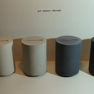 Op de afbeelding: Vier cilindrische betonnen objecten in verschillende tinten grijs en gebroken wit. Elk heeft een horizontale sleuf aan de bovenkant. De tekst "art-beton-design" staat boven de objecten.