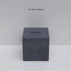 Peut inclure: Une boîte en béton gris foncé avec une fente sur le dessus. La boîte est posée sur une surface blanche. Le texte "art-beton-design" est imprimé sur une surface blanche au-dessus de la boîte.