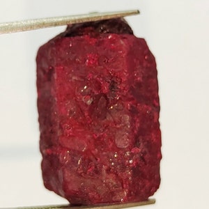 May include: A deep red, rough-cut gemstone with a slightly rectangular shape. The gemstone has a natural, unpolished finish and is a rich, vibrant red color.