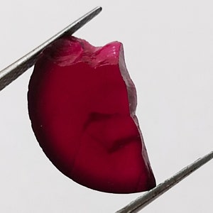 May include: A deep crimson gemstone, held by silver tweezers against a white backdrop. The gem has a smooth, polished surface with an irregular shape, highlighting its natural form. The colour is rich and saturated, with a hint of translucency.
