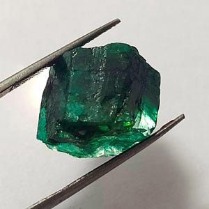 May include: A large, dark green emerald gemstone with a rough, faceted surface. The emerald is held in tweezers against a white background.